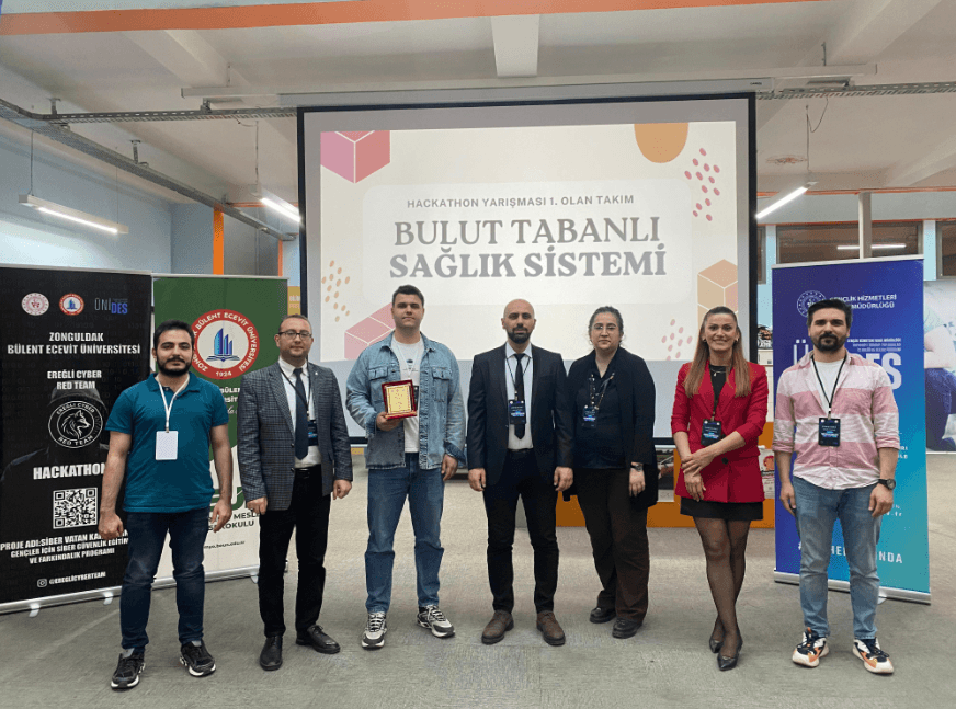Hackathon ödül töreninde birincilik plaketiyle çekilmiş grup fotoğrafı. Arkadaki ekranda “Hackathon yarışması 1. olan takım” yazıyor.